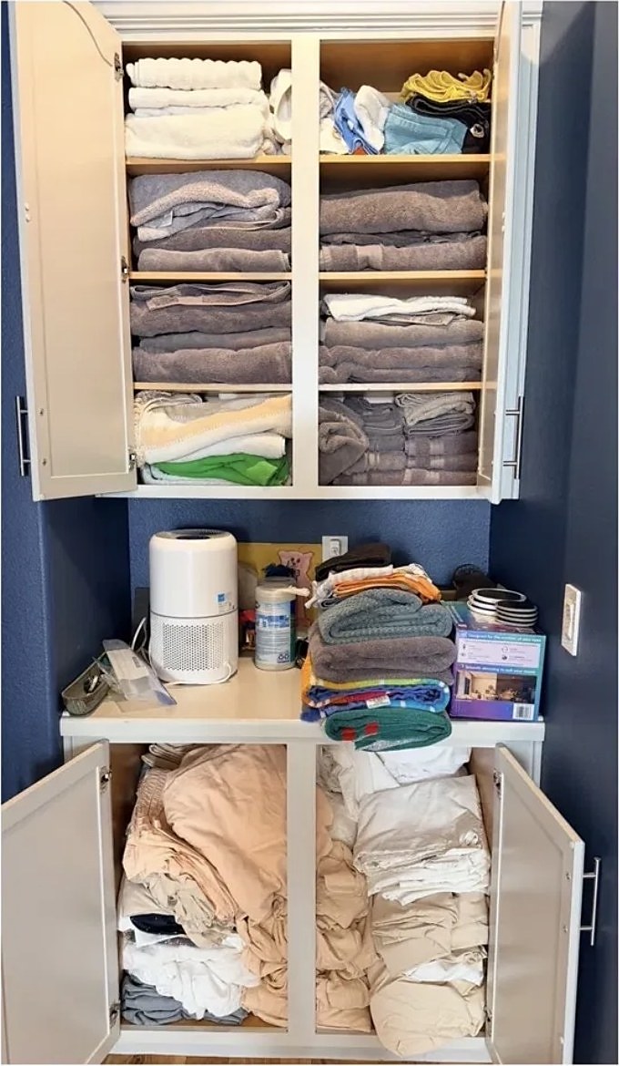 Linen cabinet with piles of unfolded towels and bedding before organizing